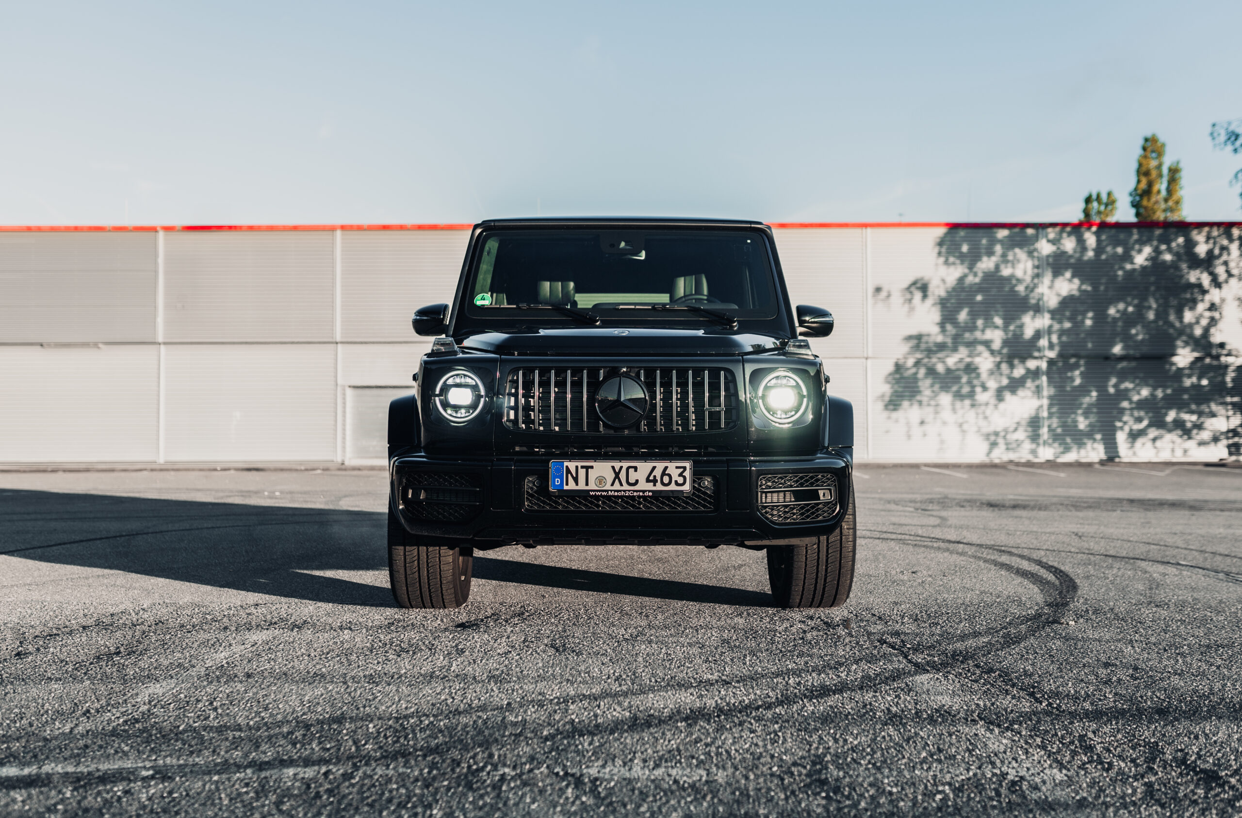 Schwarzer Mercedes-Benz G-Klasse, frontal, Scheinwerfer an, auf leerem Parkplatz bei Tageslicht.